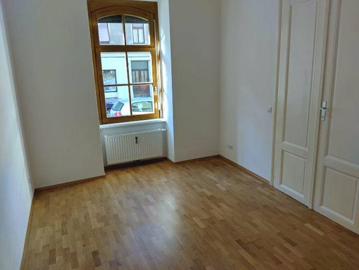 Helles Zimmer mit Parkettboden, weißer Tür und einem großen Fenster mit Blick auf die Straße.