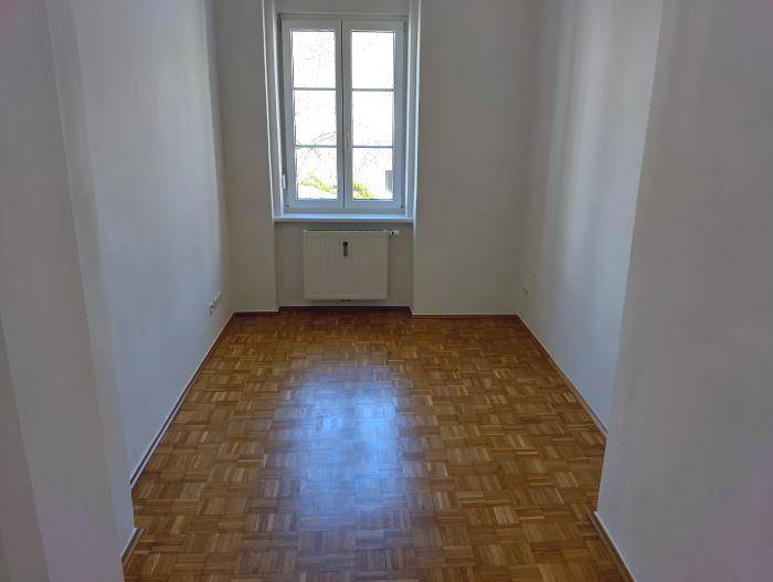 Kleineres Zimmer mit Würfelparkett, einem Fenster und einem Heizkörper.