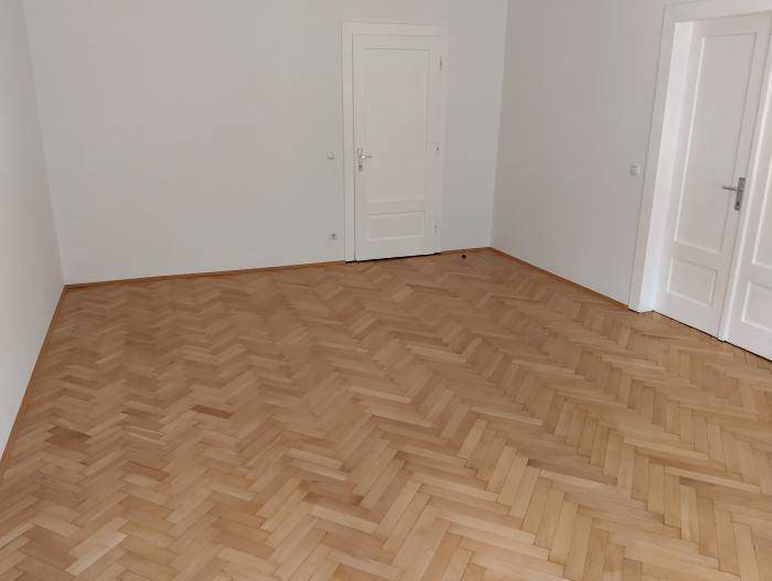 Leeres Zimmer mit hellem Fischgrätparkett und zwei weißen Türen in gutem Zustand.