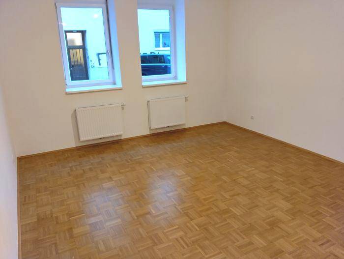 Heller Raum mit Parkettboden, zwei Fenstern und Heizkörpern, Blick auf die Straße.
