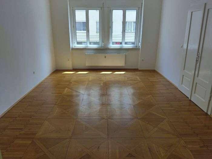 Heller, unmöblierter Raum mit klassischem Parkettboden und großen Fenstern, die viel Licht hereinlassen.