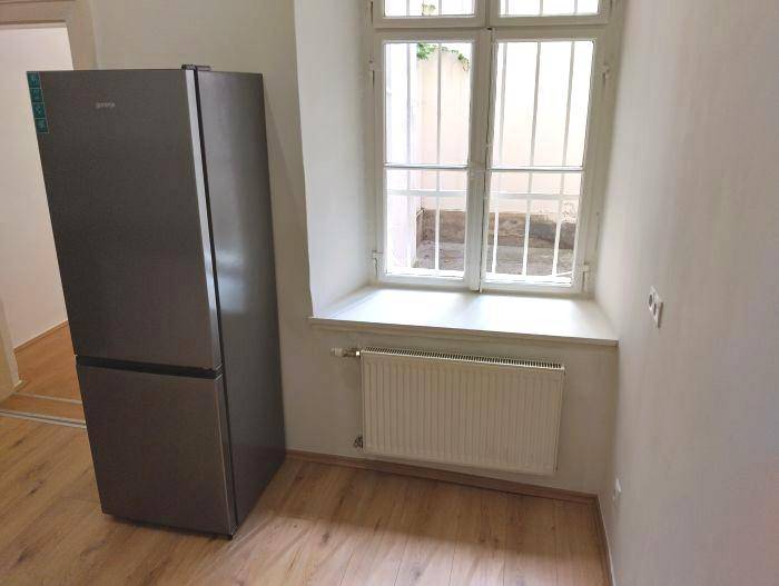 Bereich mit großem Kühlschrank, Fenster mit Gitter und Heizkörper, auf Holzfußboden.