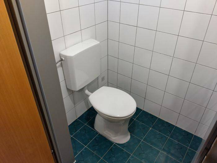 Separates WC mit weißer Toilette und grünen Bodenfliesen, sauber und funktional gestaltet.
