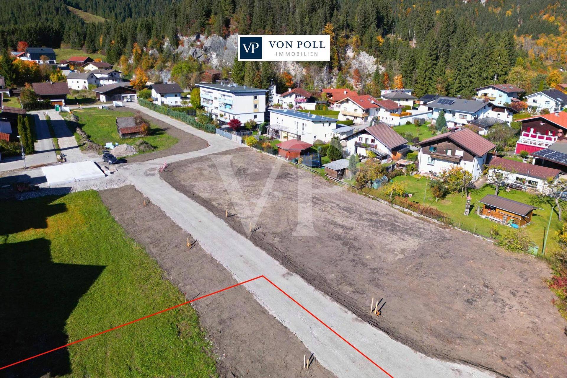 Luftaufnahme eines Baugrundstücks in einer Siedlung mit Blick auf bewaldete Berge im Hintergrund.
