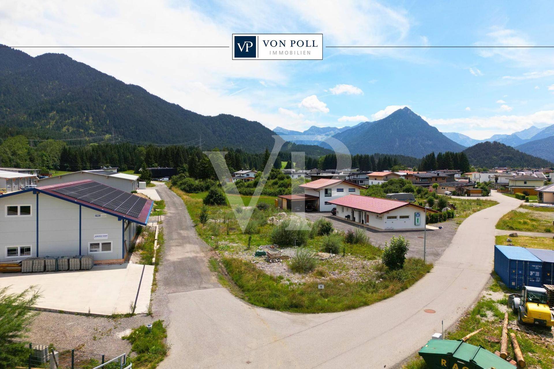 Panoramablick auf eine ländliche Gegend mit Gewerbegebäuden, unbebauten Flächen und majestätischen Bergen im Hintergrund.