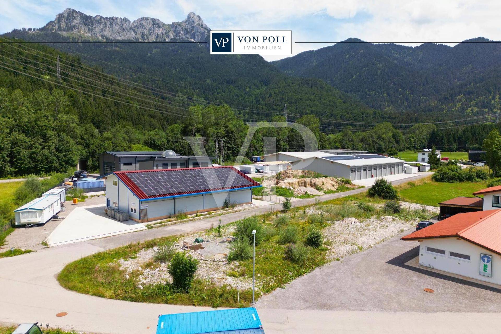 Luftaufnahme einer Gewerbezone mit mehreren Gebäuden, Solaranlagen und einer beeindruckenden Bergkulisse.