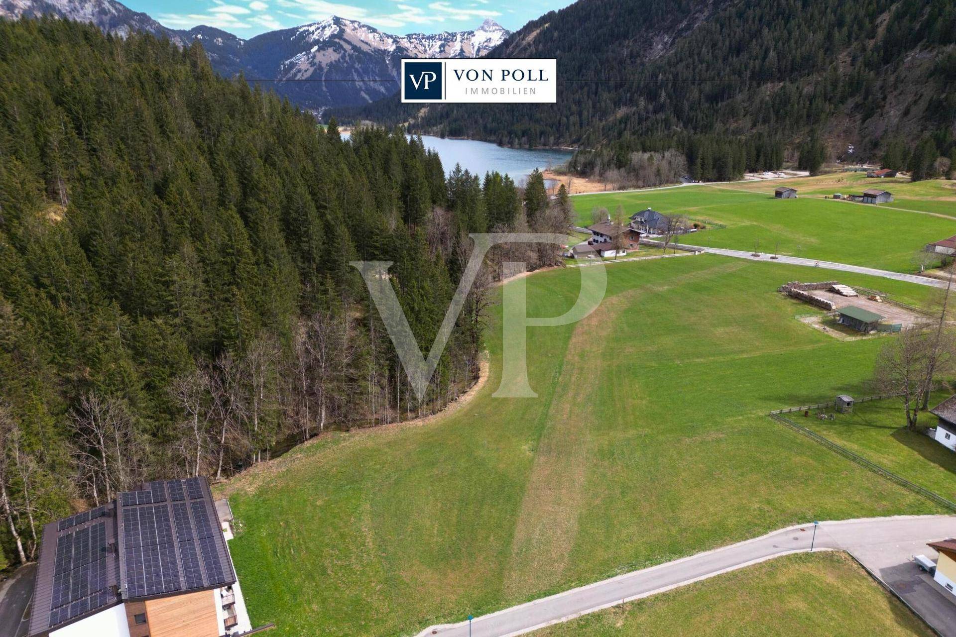 Luftaufnahme einer grünen Wiese mit Wald, Bergen und einem See, im Vordergrund ein Gebäude mit Solaranlage.