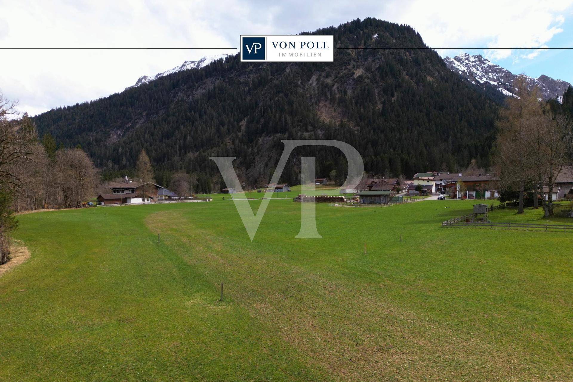 Weitläufige grüne Wiesenfläche mit traditionellen Häusern im Hintergrund, umgeben von Bergen und Wäldern.