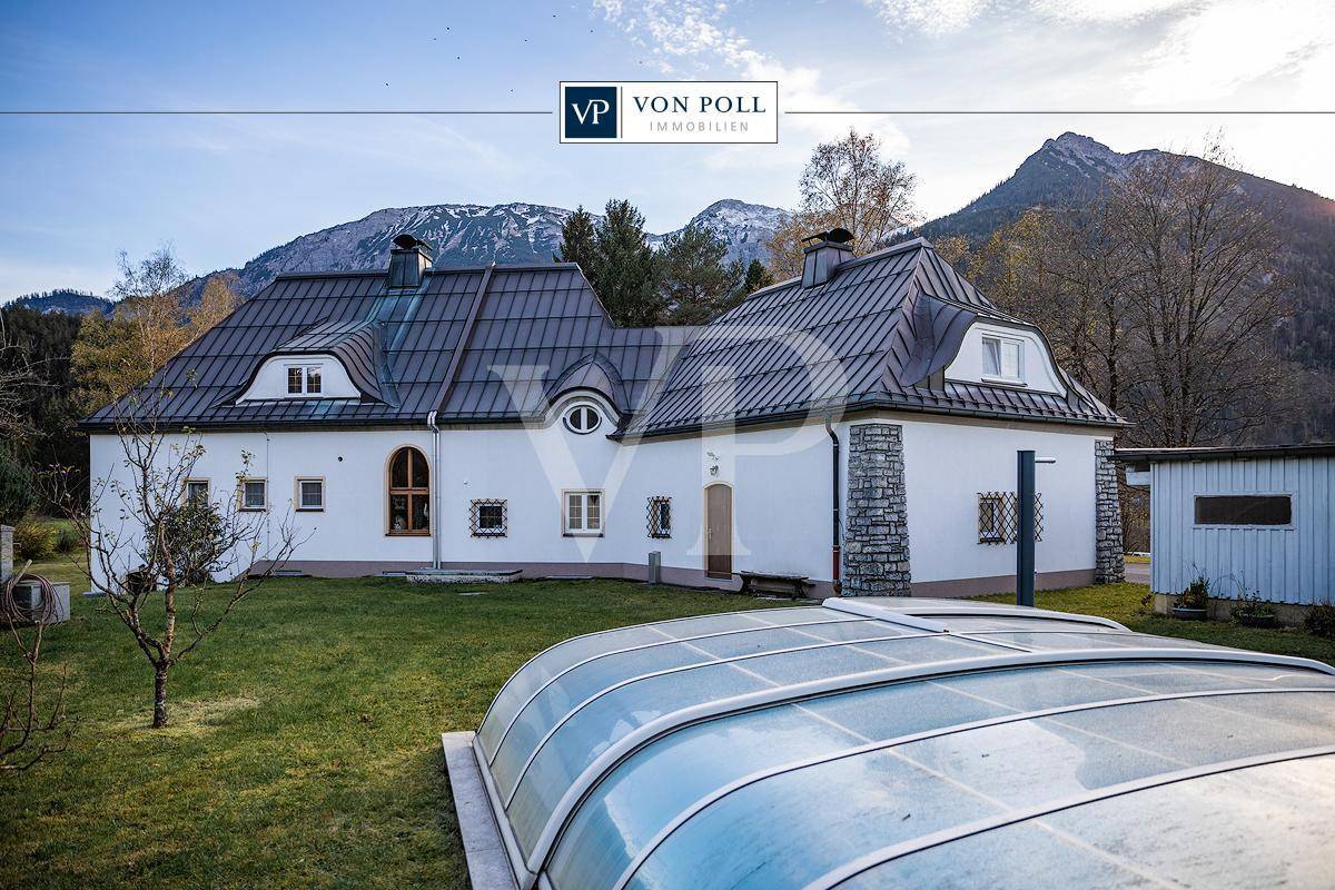 Landhaus mit weißer Fassade, großem Garten und überdachtem Pool vor beeindruckender Bergkulisse.