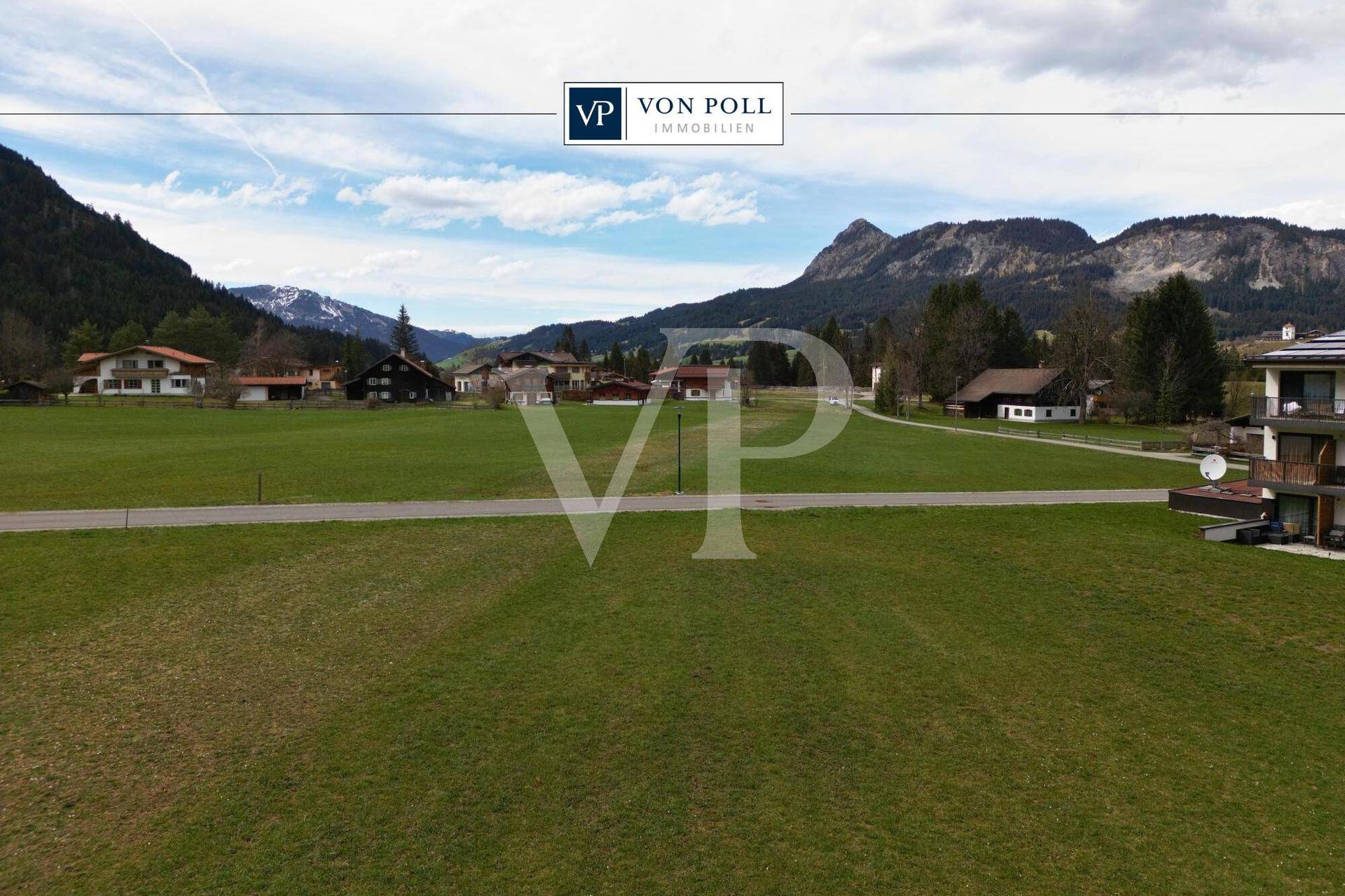 Weitläufige grüne Wiesenfläche mit einer Straße, umgeben von traditionellen Häusern und majestätischen Bergen.