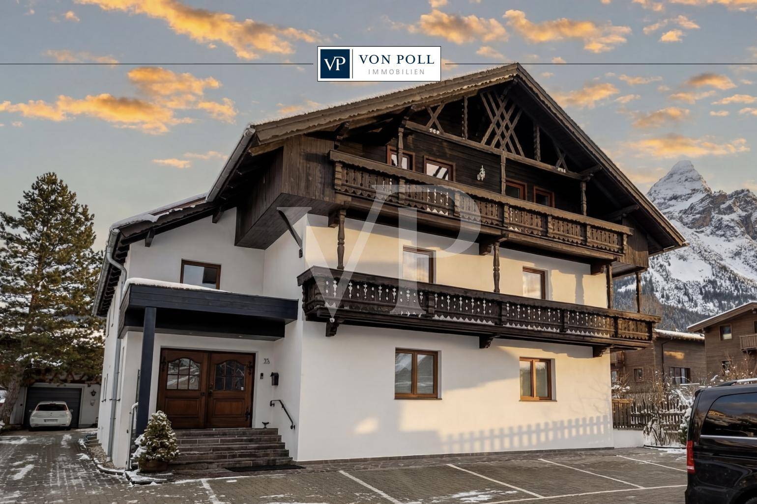 Traditionelles Chalet-Haus mit Holzbalkonen und schneebedeckten Bergen im Hintergrund bei Sonnenuntergang.