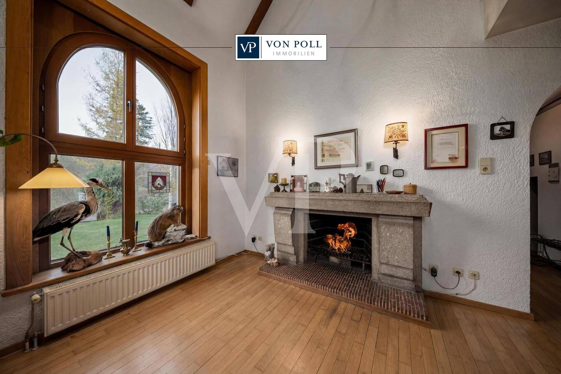 Gemütliches Wohnzimmer mit Holzboden, großem Kamin und hohen Decken, umgeben von natürlichem Licht.