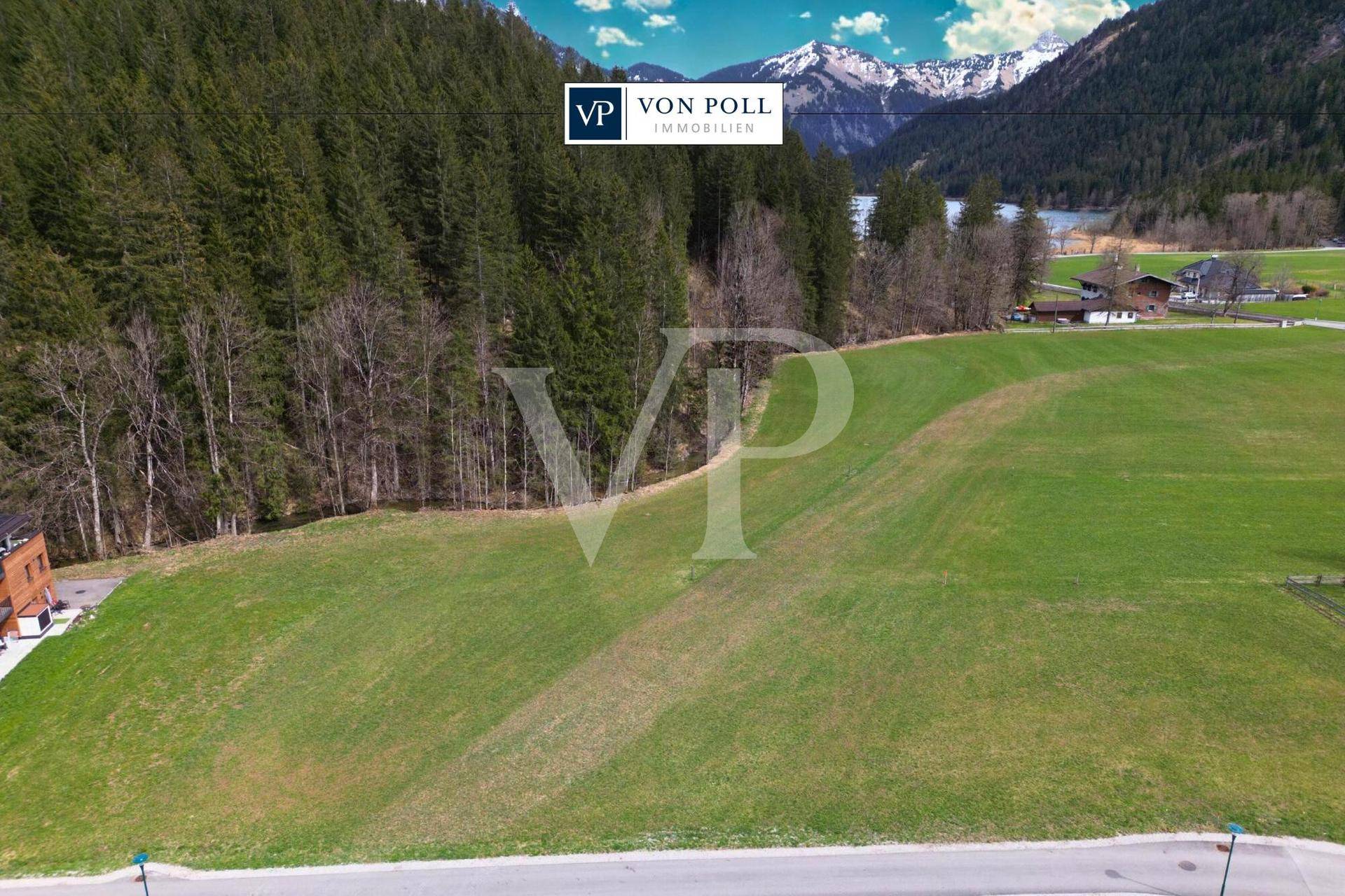 Weitläufige grüne Wiesenfläche mit angrenzendem Wald, Bergen und einem See im Hintergrund unter blauem Himmel.