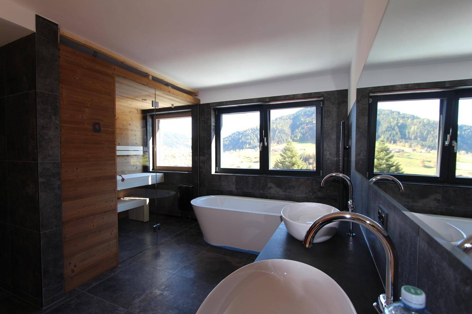 Modernes Badezimmer mit freistehender Badewanne, dunklen Fliesen und Panoramablick auf die Berge.