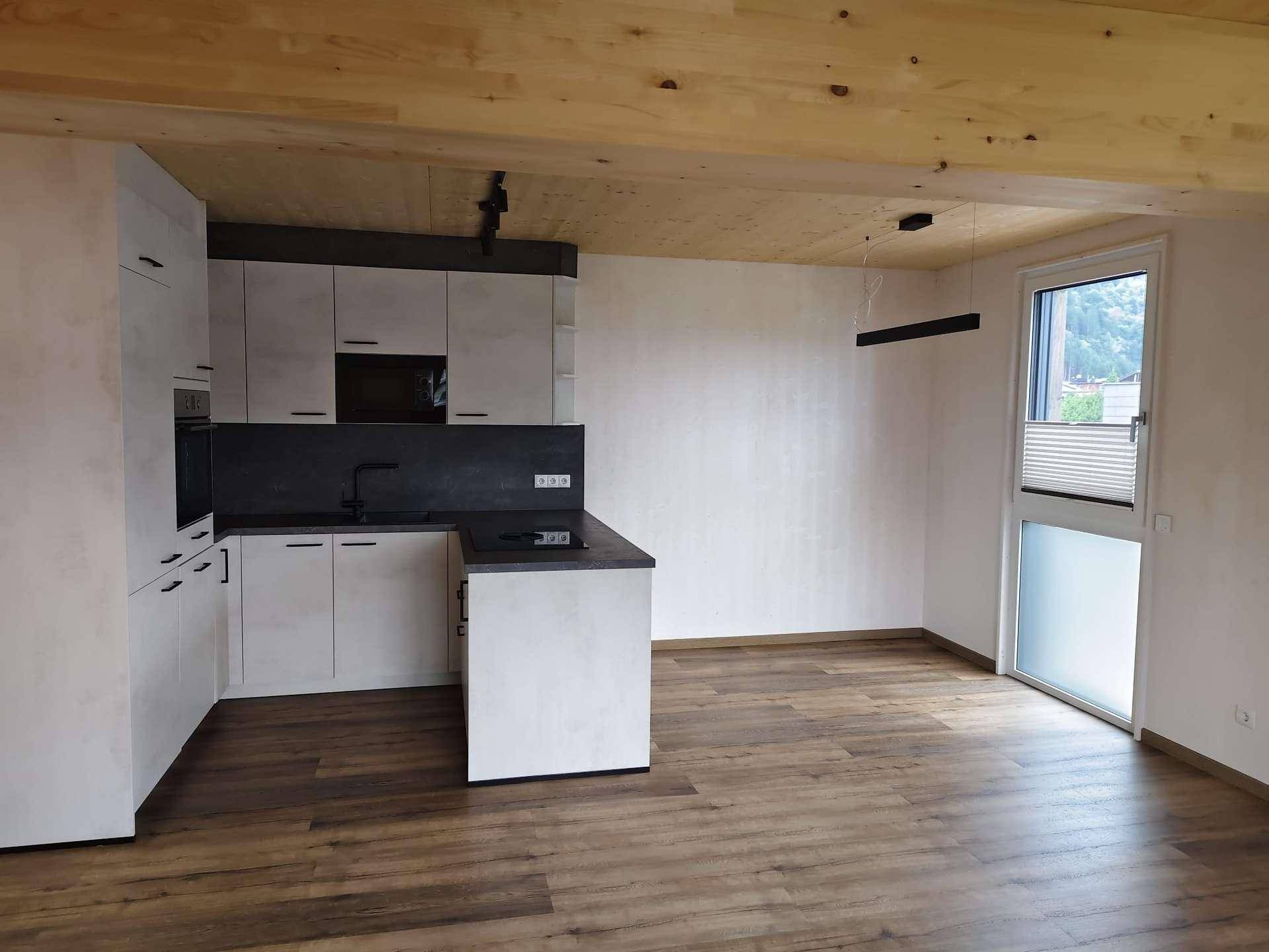Heller Küchenbereich mit moderner Einbauküche, Holzbalkendecke und Zugang zu einem Fenster mit Jalousie.