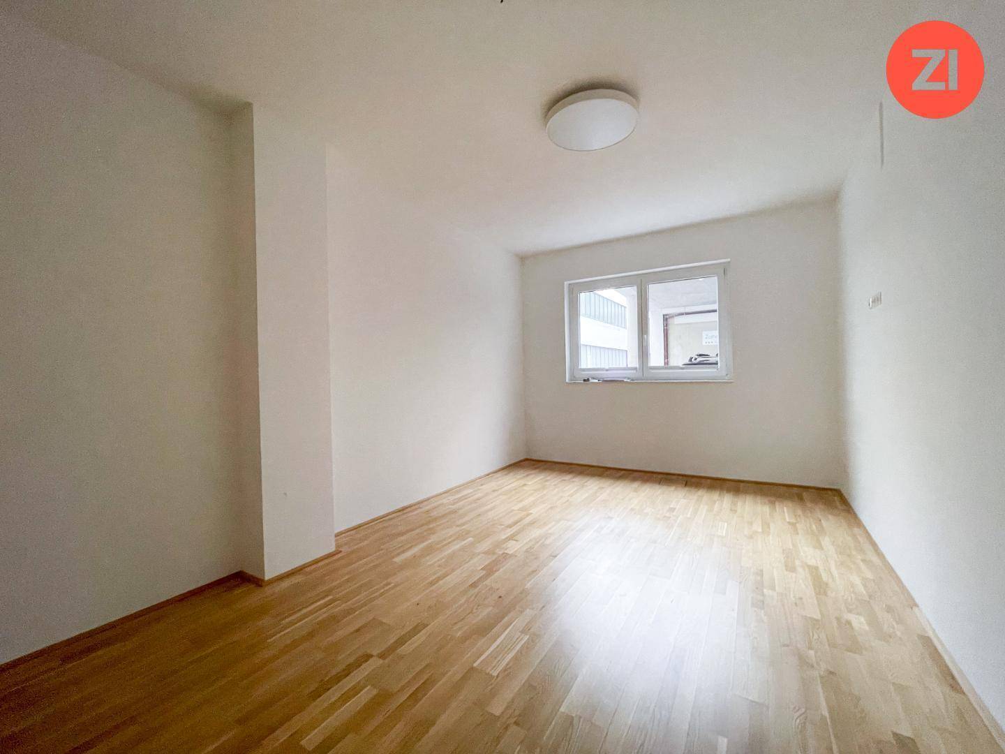 Kleiner, leerer Büroraum mit Holzboden, weißen Wänden und einem Fenster mit Blick nach draußen.