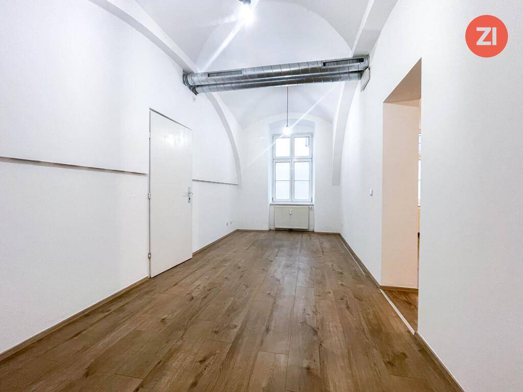 Leerer Raum mit Holzboden, weißen Wänden, einem Fenster und einer markanten Gewölbedecke.