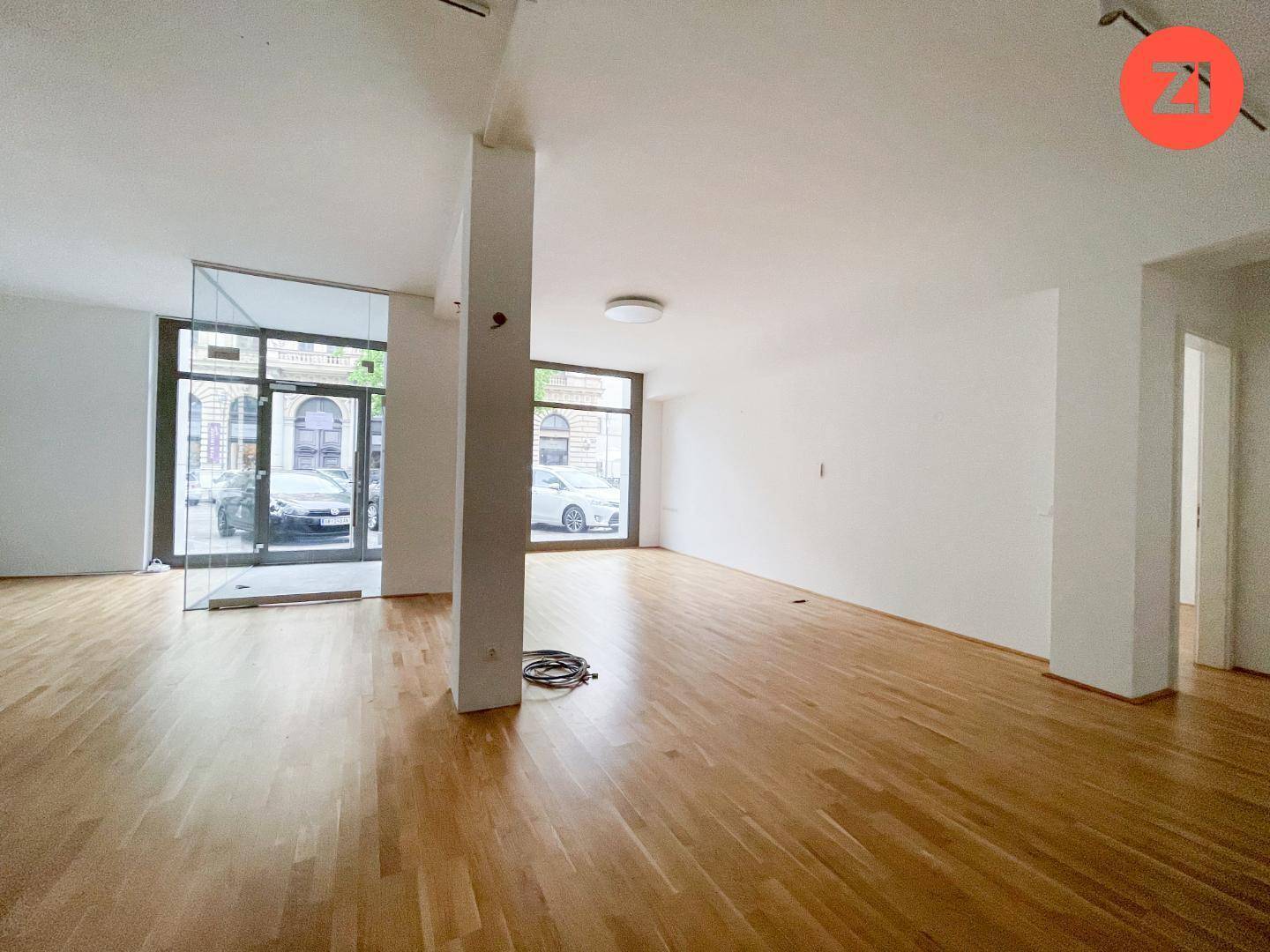Heller Gewerberaum mit Holzboden, großen Fenstern zur Straße und einer modernen Glasbox im Vordergrund.