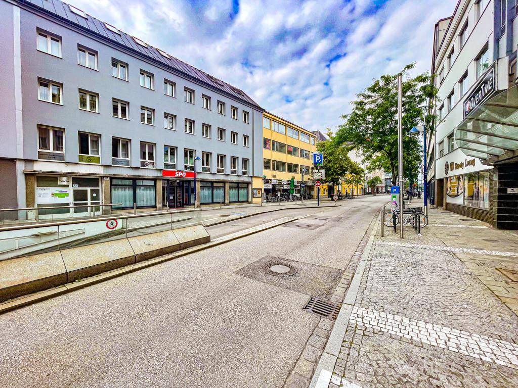 Blick auf eine städtische Straße mit Gebäuden, Geschäften und parkenden Fahrrädern unter bewölktem Himmel.