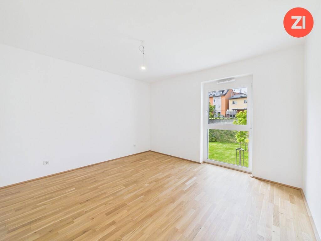 Leeres Zimmer mit Holzboden und großem Fenster, das einen Blick auf den grünen Garten bietet.