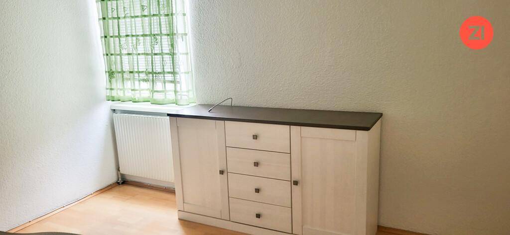 Helles Zimmer mit Fenster, Heizkörper und einem weißen Sideboard auf Holzfußboden.