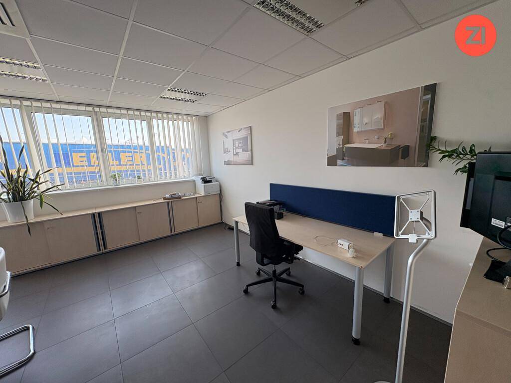 Helles Büro mit Schreibtisch, Bürostuhl und Fenstern mit Jalousien und Blick nach draußen.