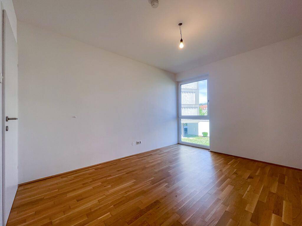 Helles Schlafzimmer mit Parkettboden und großem Fenster, das viel Tageslicht hereinlässt.