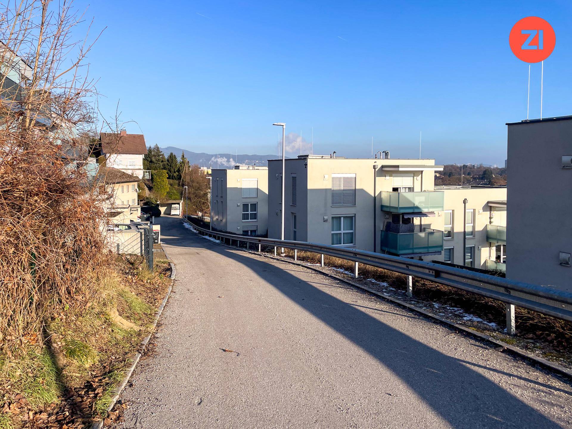 Straße führt bergauf vorbei an modernen Gebäuden mit Balkonen und einem weiten Blick in die Ferne.