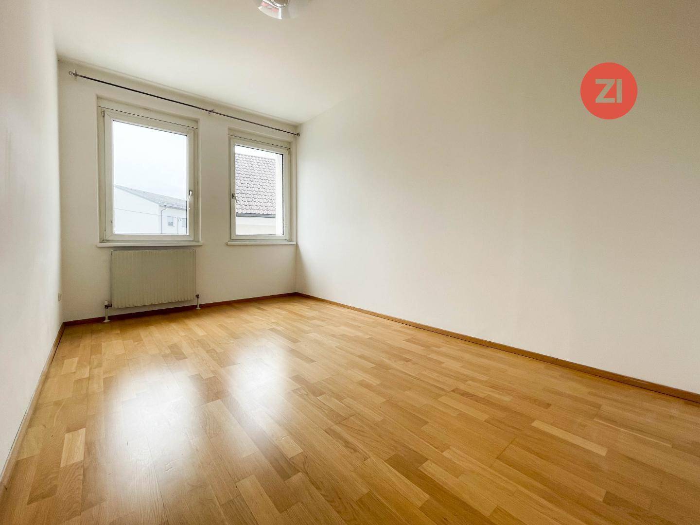 Leeres Schlafzimmer mit zwei Fenstern, Holzboden und Heizkörper, das eine angenehme Atmosphäre schafft.