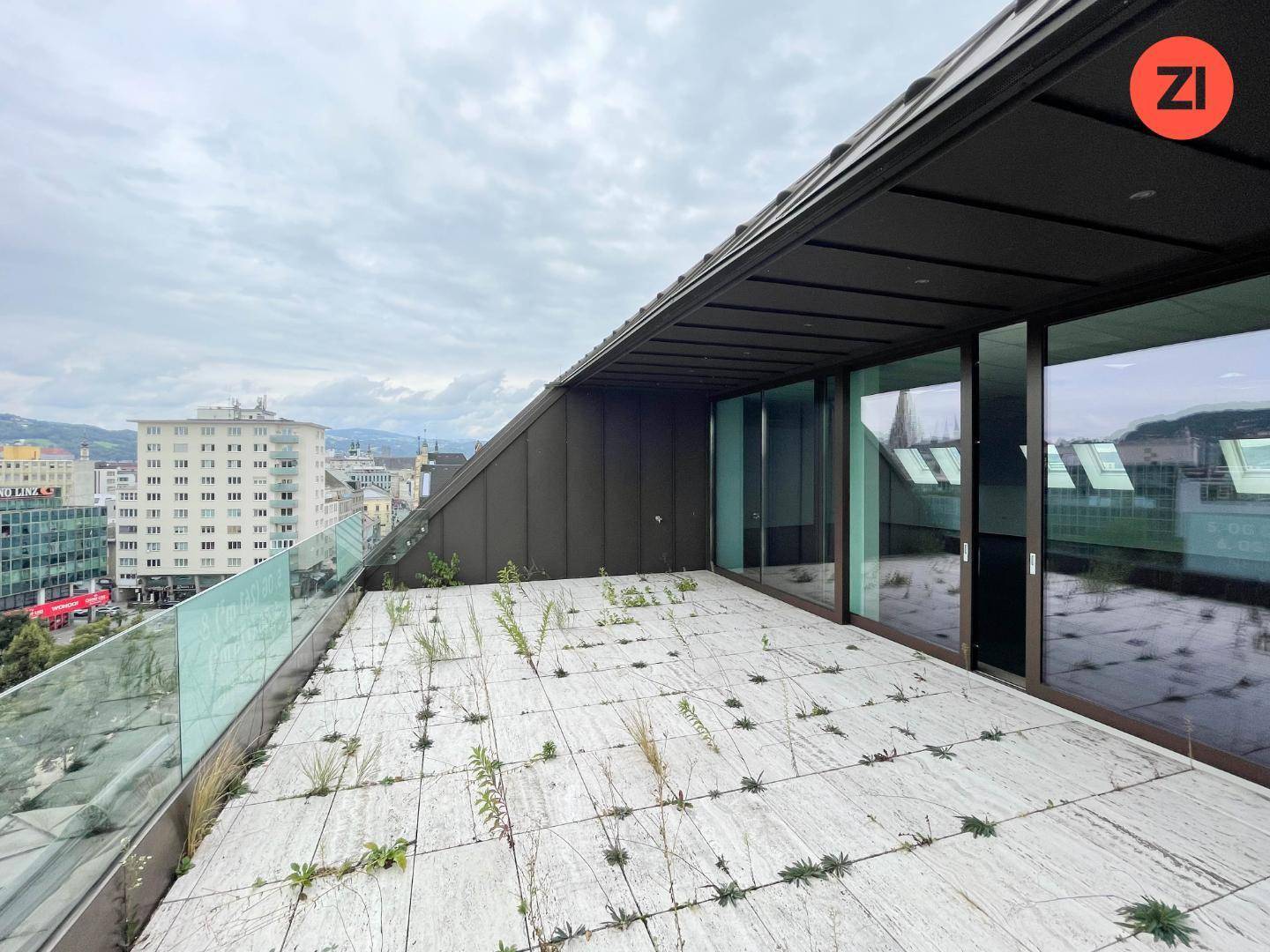 Weitläufige Dachterrasse mit Fliesenboden, Glasgeländer und Blick auf die Stadt.