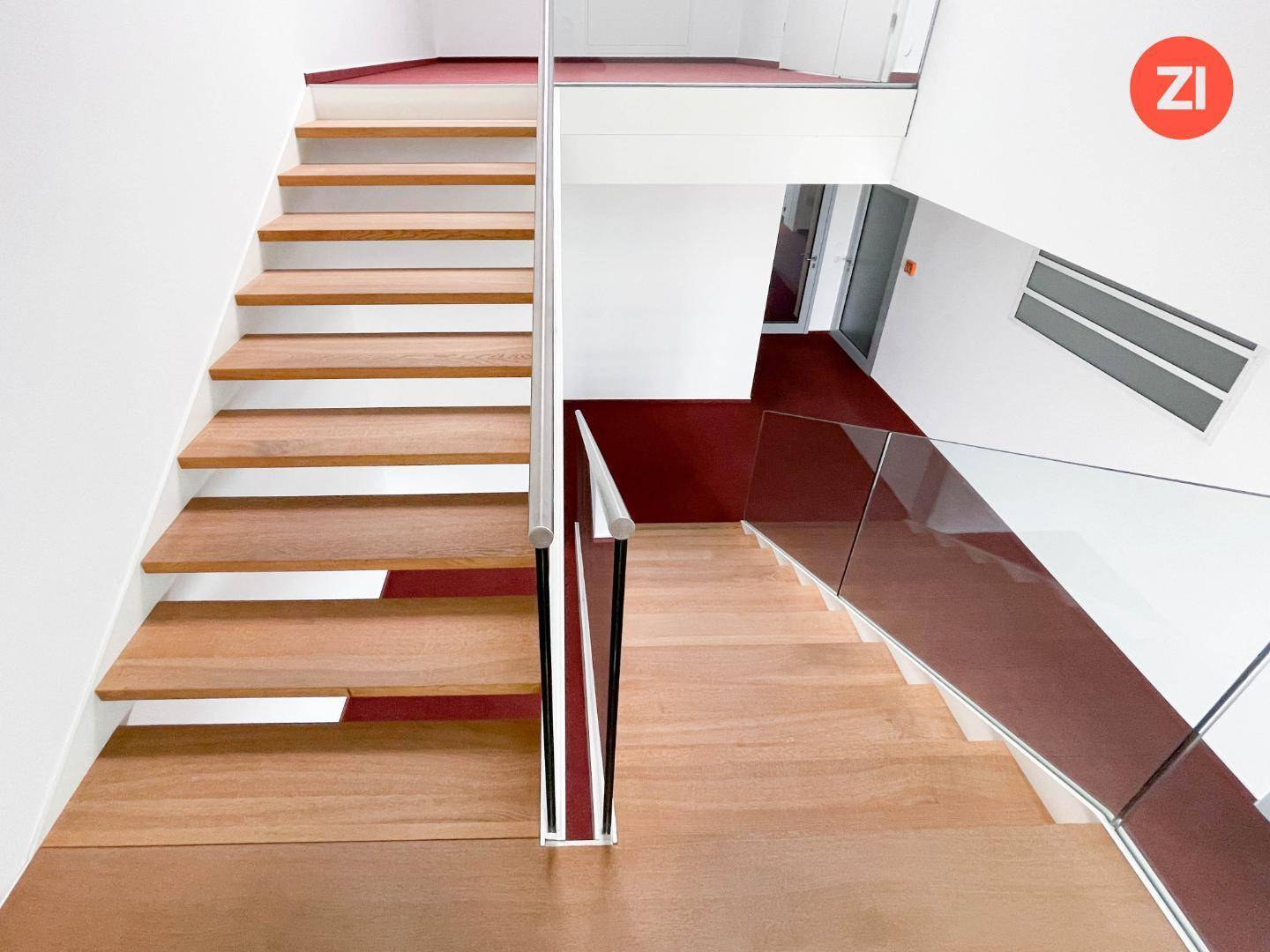 Moderne Holztreppe mit Glasgeländer und rotem Teppichboden in einem hellen Treppenhaus.