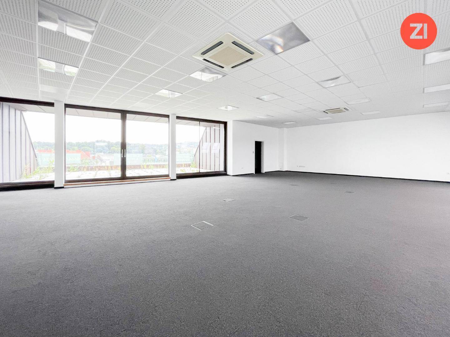 Geräumiger, leerer Büroraum mit Teppichboden, großen Fenstern und abgehängter Decke.