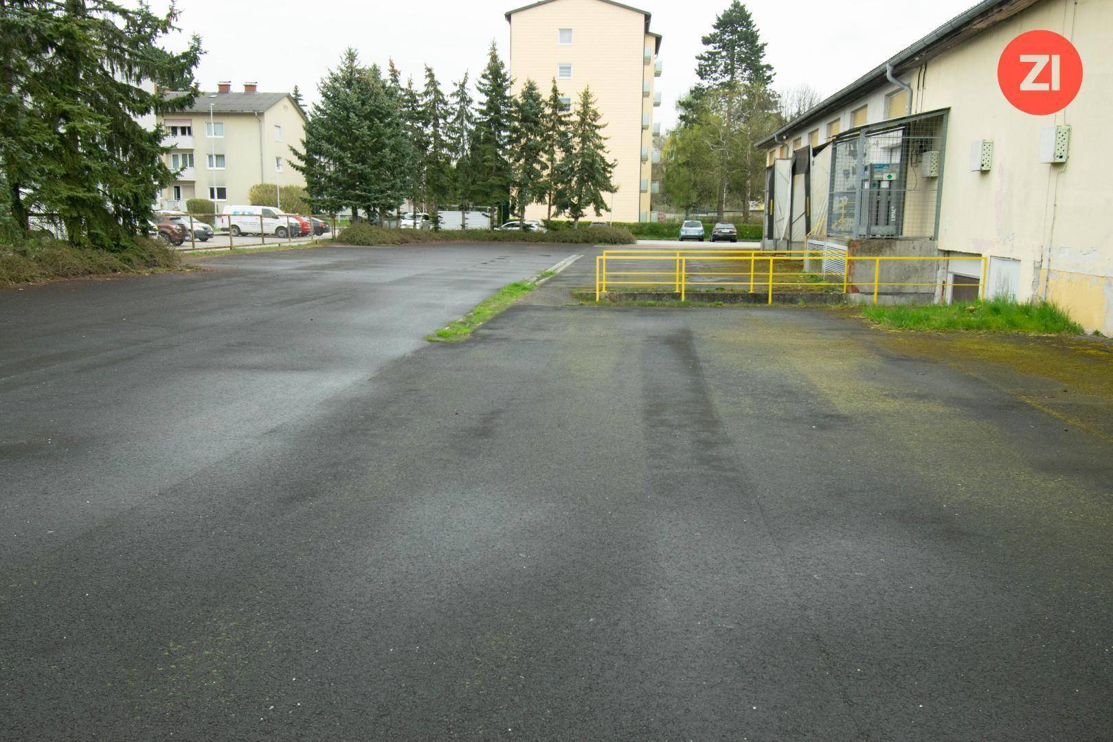 Großer asphaltierter Parkplatz vor einem Gebäude mit gelben Geländern und Bäumen im Hintergrund.