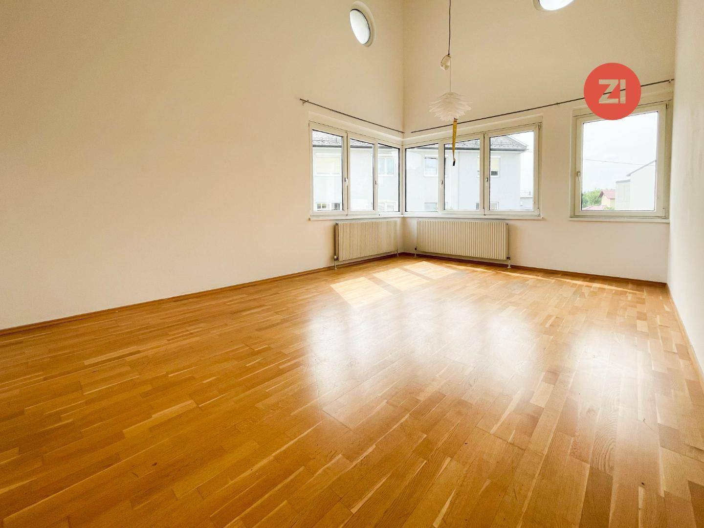 Geräumiger, leerer Wohnraum mit Holzboden und vielen Fenstern, die für Helligkeit sorgen.