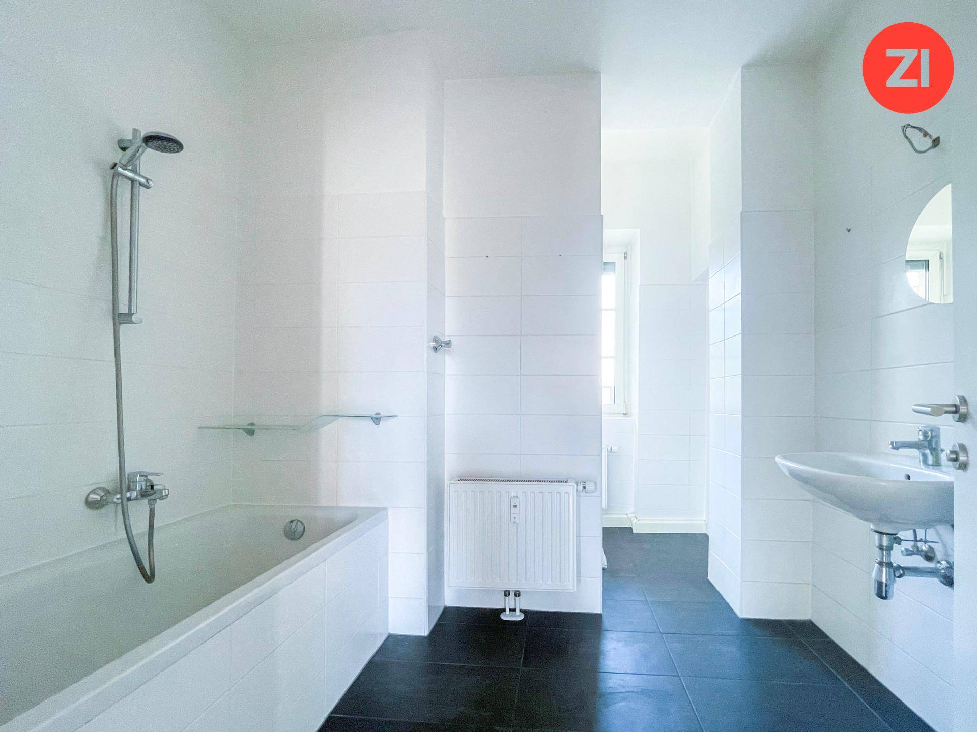 Funktionales Badezimmer mit Badewanne, Duscharmatur, Waschbecken und dunklen Bodenfliesen.