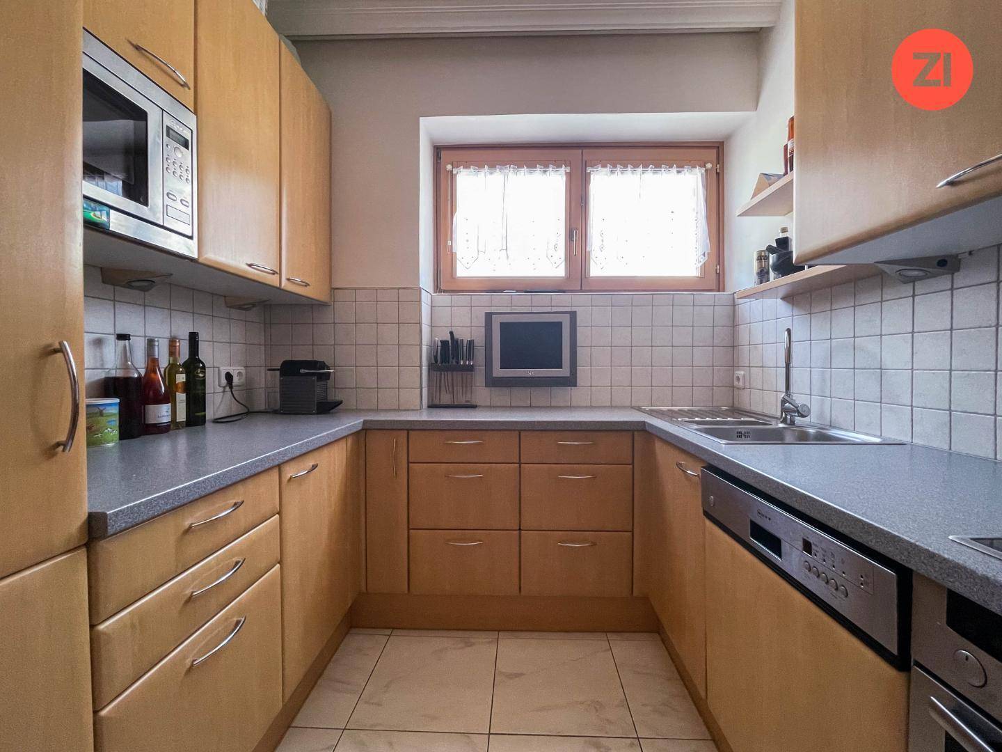 Funktionale Küche mit hellen Holzfronten, Fliesenboden und Fenster über der Spüle.
