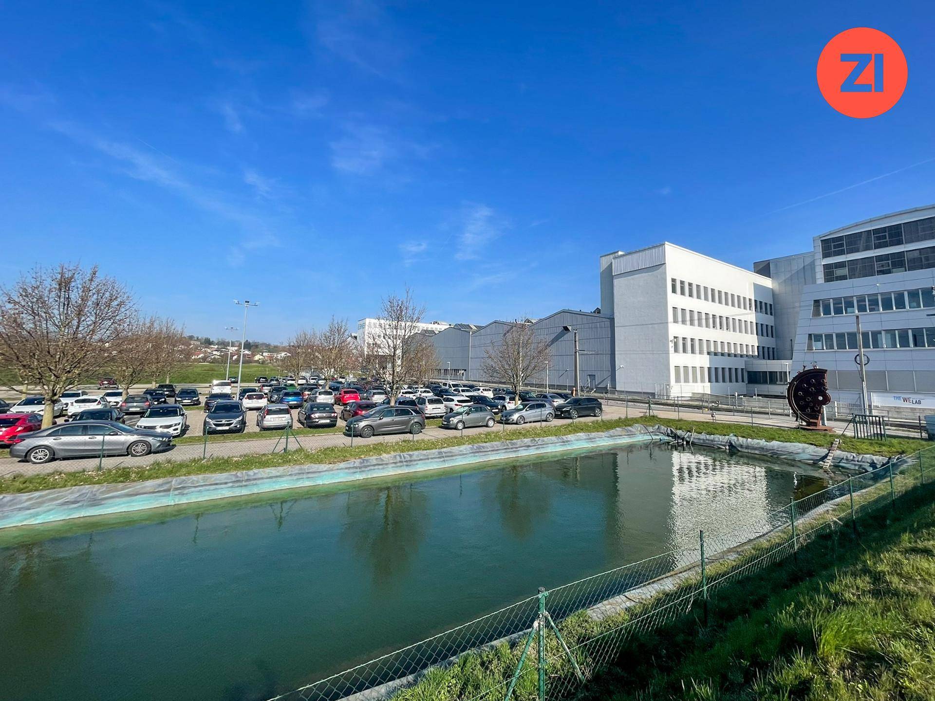 Außenansicht eines Industriegebäudes mit großem Parkplatz und einem Teich im Vordergrund unter blauem Himmel.