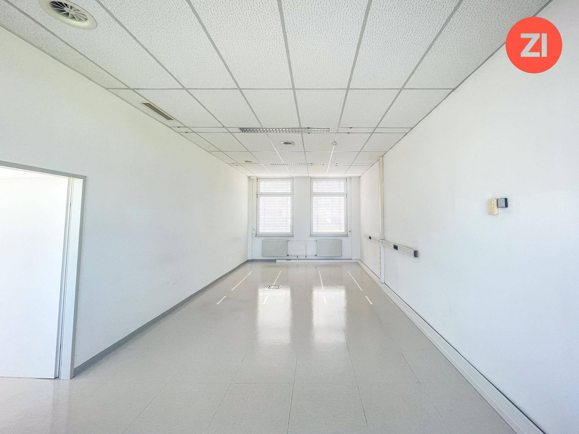 Kleinerer, leerer Büroraum mit zwei Fenstern, abgehängter Decke und weißen Wänden.