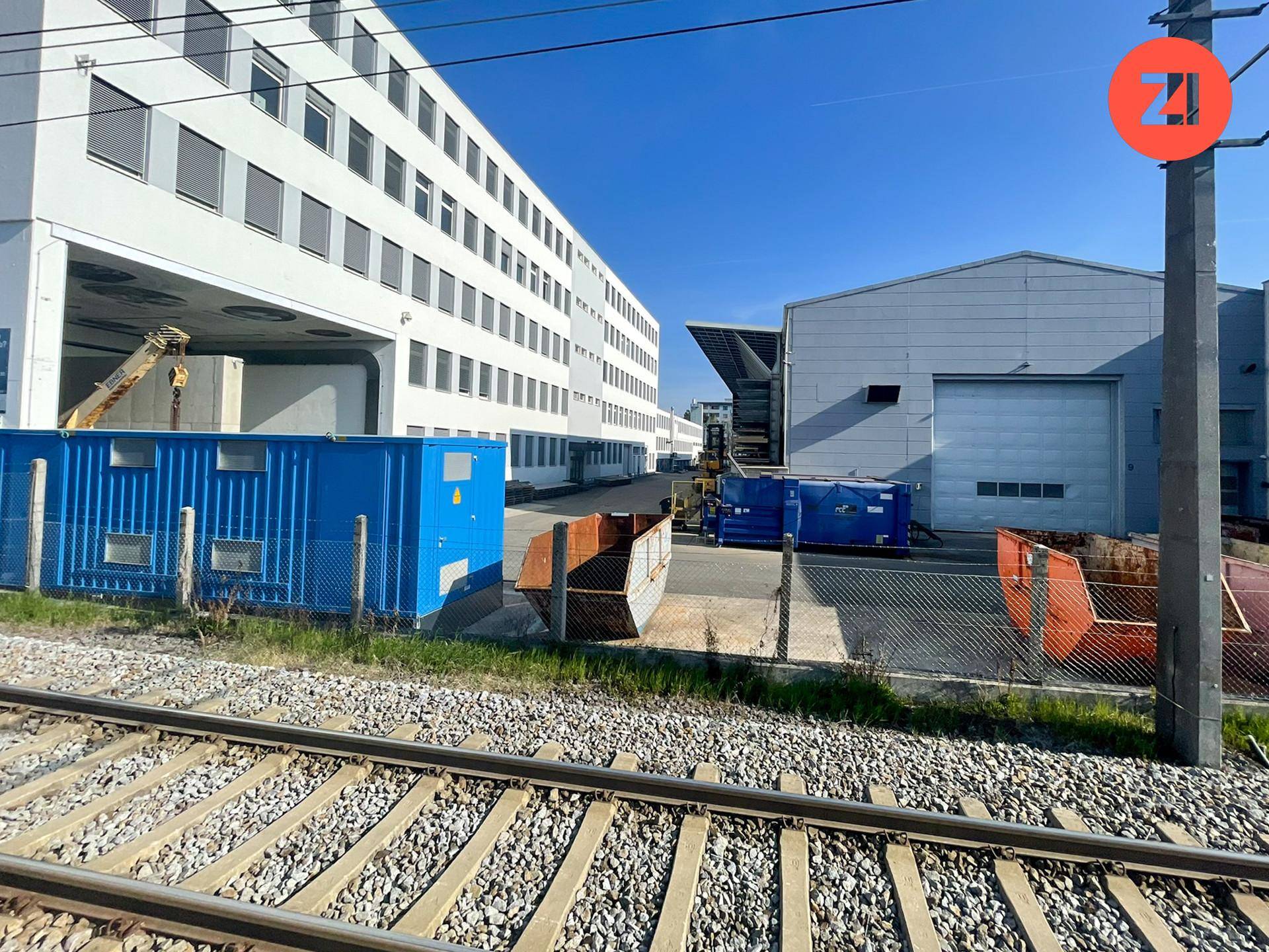 Außenansicht von zwei Industriegebäuden mit Bahngleisen, blauen Containern und Abfallmulden im Vordergrund.