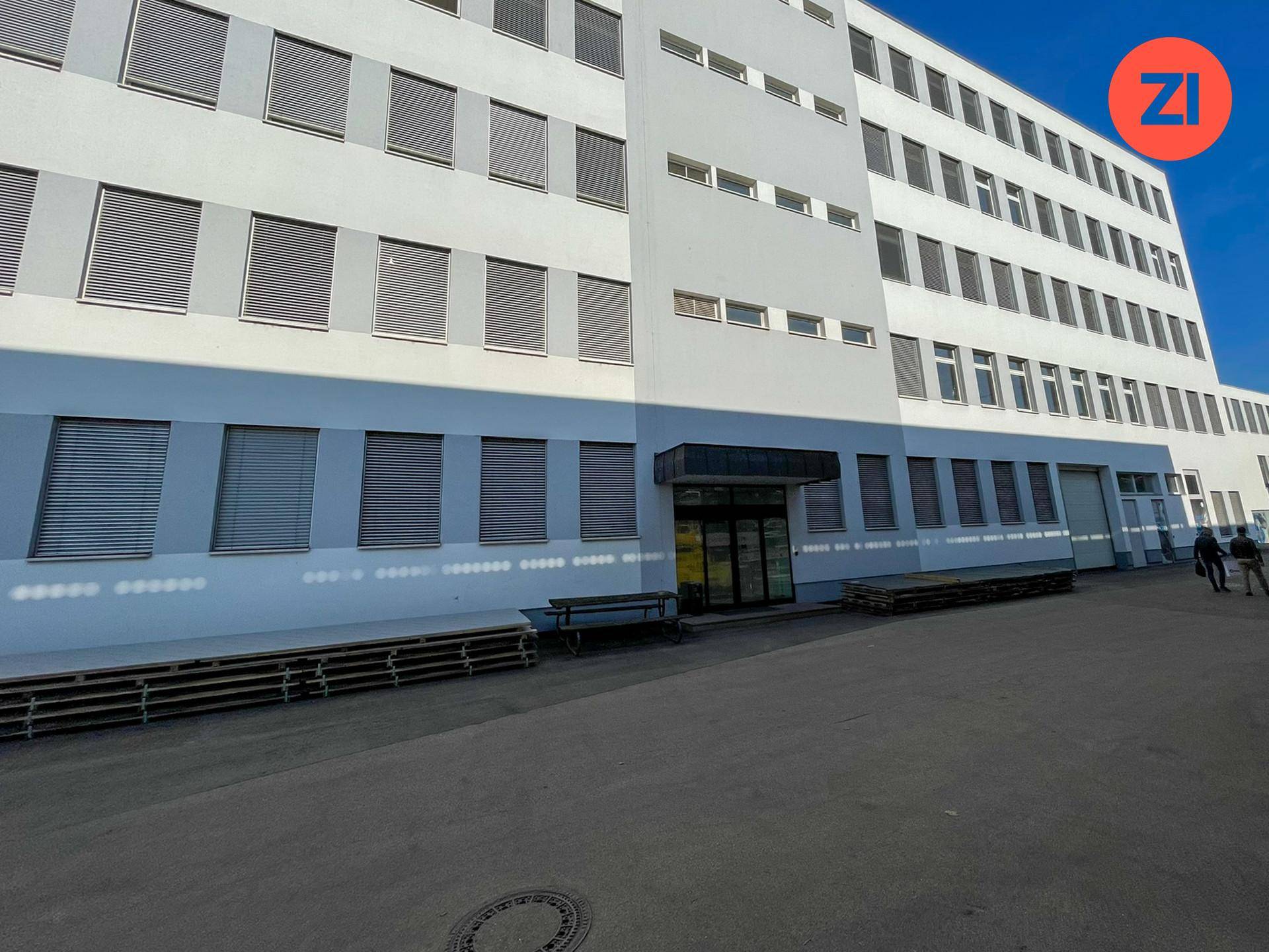 Eingangsbereich eines modernen weißen Gebäudes mit vielen Fenstern und Paletten vor der Fassade.