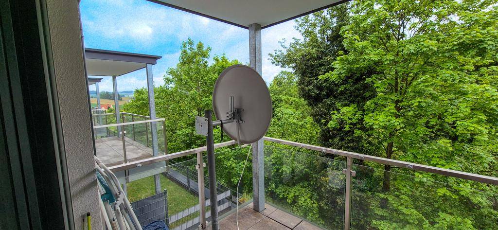Balkon mit Glasgeländer und Satellitenschüssel, bietet einen weiten Blick ins Grüne.