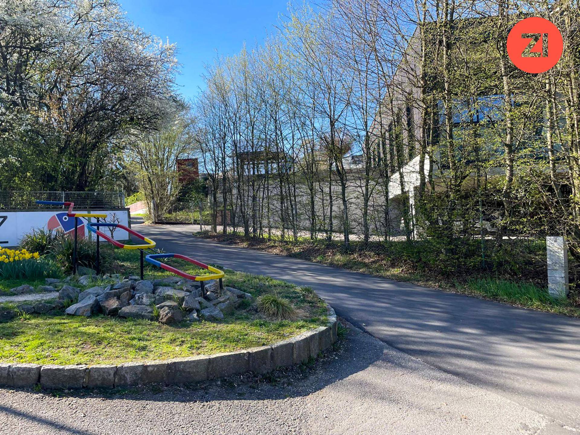 Zufahrtsweg zu einem Gebäude mit einem kleinen Spielbereich und Bäumen im Hintergrund.