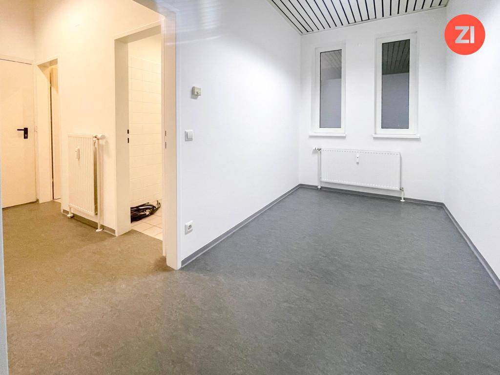 Kleinerer Büroraum mit zwei Fenstern, Heizkörper und Zugang zu einem gefliesten Bereich.