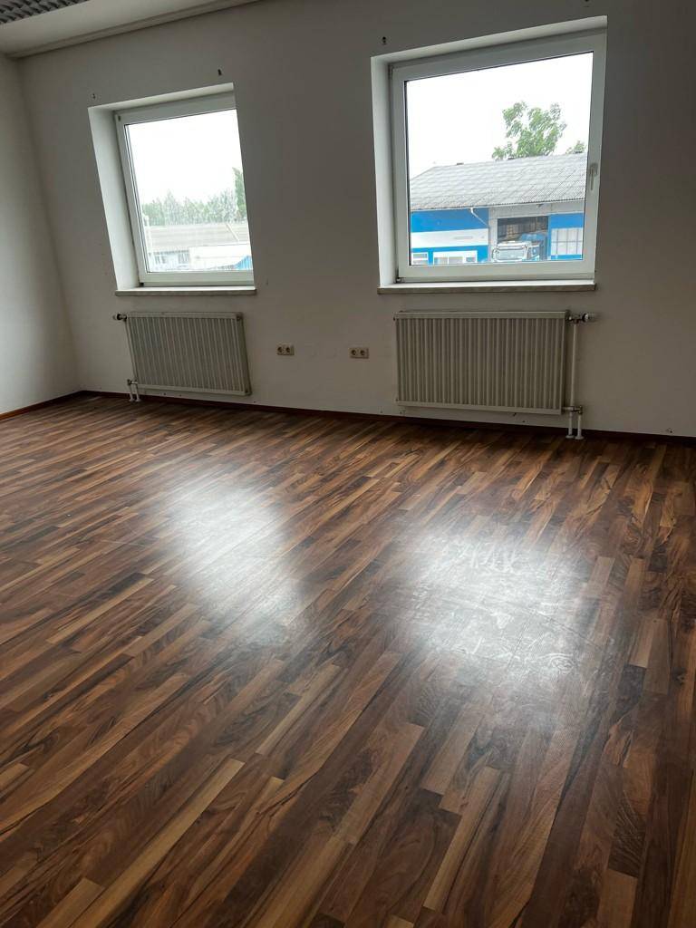 Heller Büroraum mit zwei Fenstern, Heizkörpern und glänzendem Laminatboden, der das Licht reflektiert.