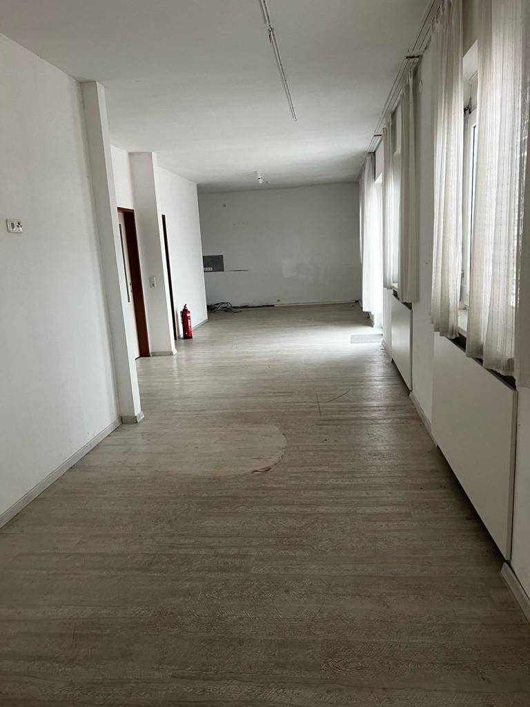 Langer Flur mit hellem Fliesenboden, Fenstern mit Vorhängen und einem Feuerlöscher an der Wand.