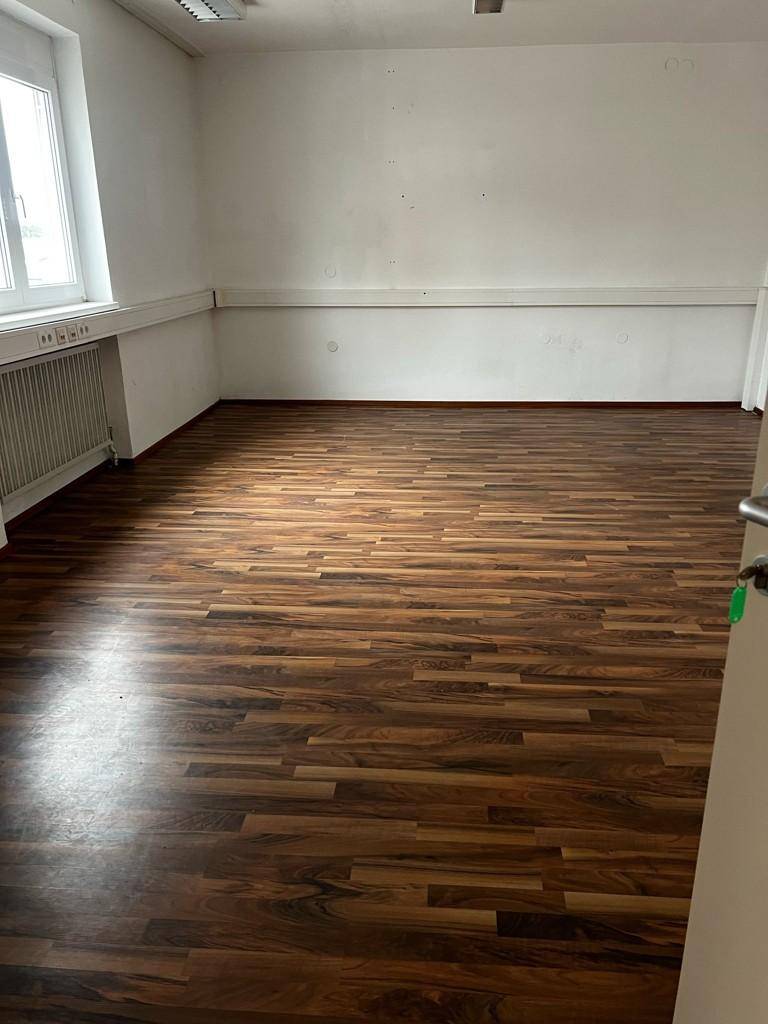 Leerer Büroraum mit Laminatboden, einem Fenster und einer weißen Wand, die auf Renovierungsbedarf hindeutet.