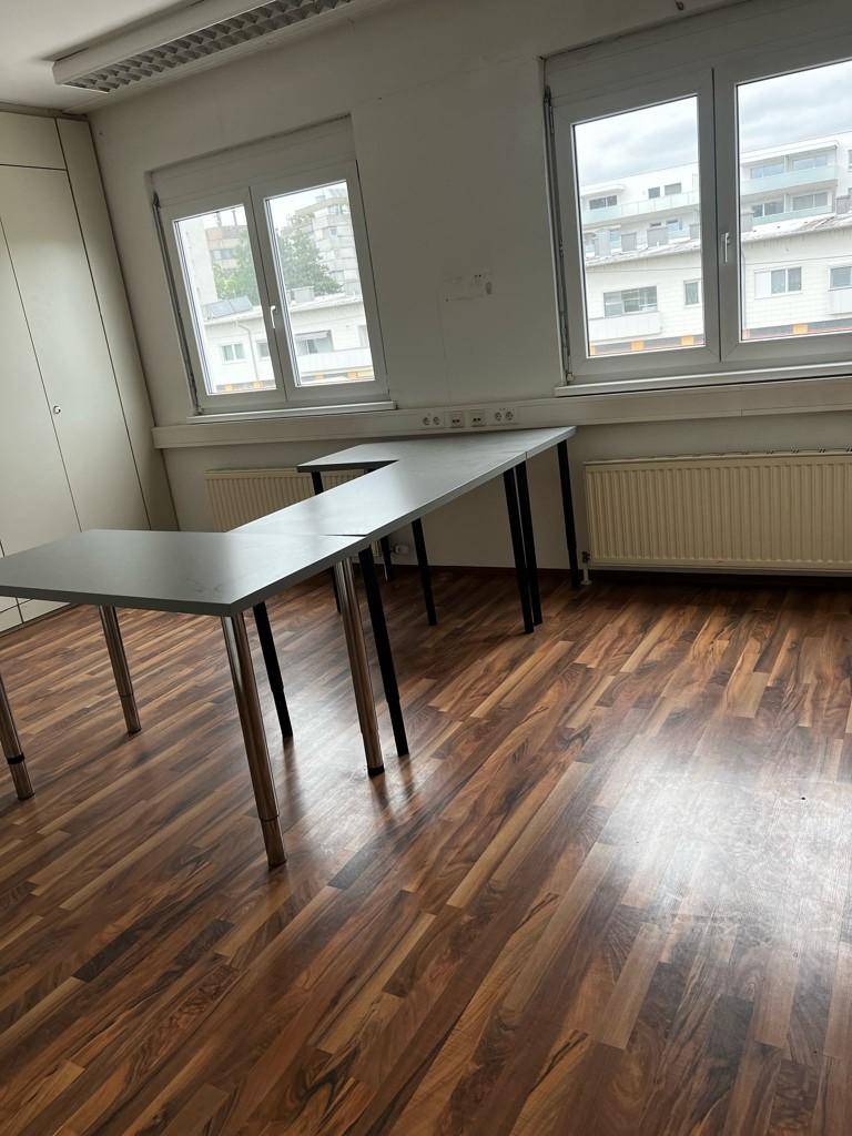 Büroraum mit Laminatboden, zwei Fenstern und einem L-förmigen Schreibtisch, der den Raum strukturiert.