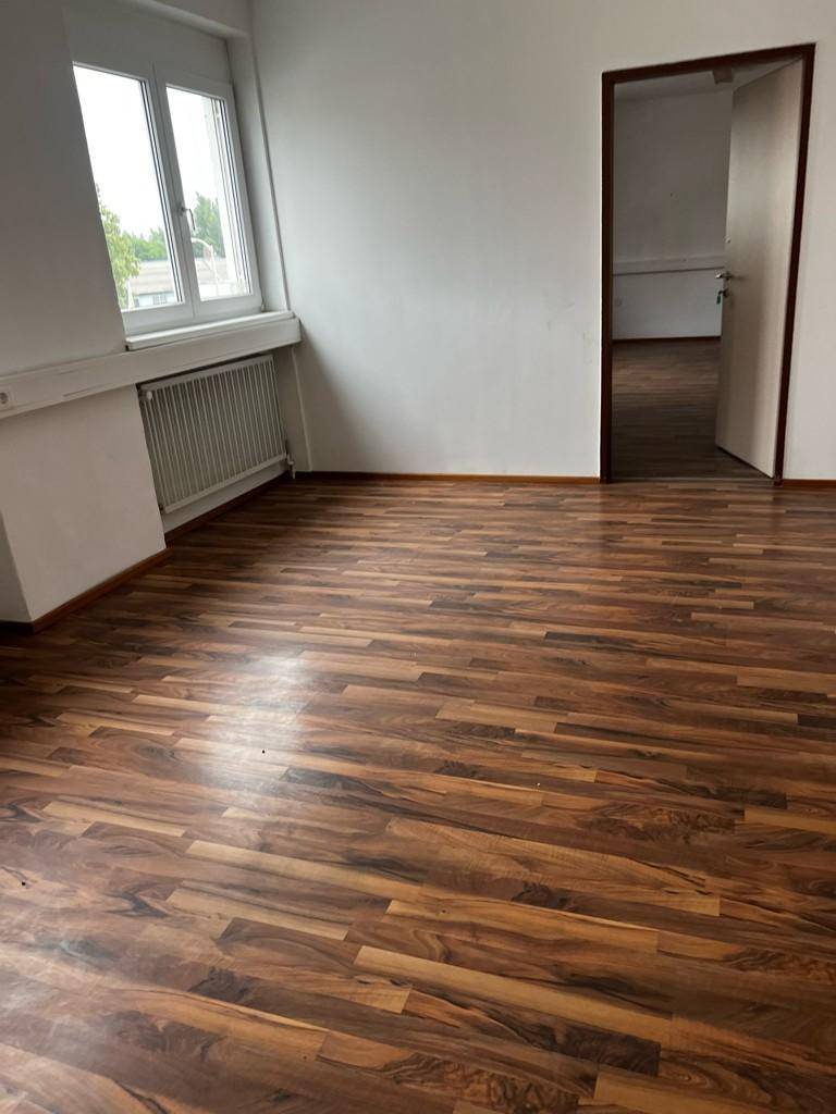 Heller Büroraum mit Laminatboden und einem Fenster, das den Blick nach draußen freigibt.