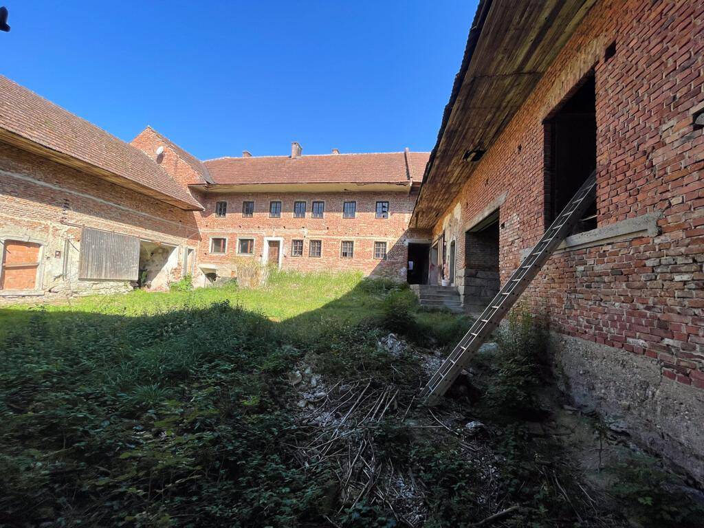 Innenhof des Bauernhofkomplexes mit Ziegelgebäuden, Fenstern und überwuchertem Gras.