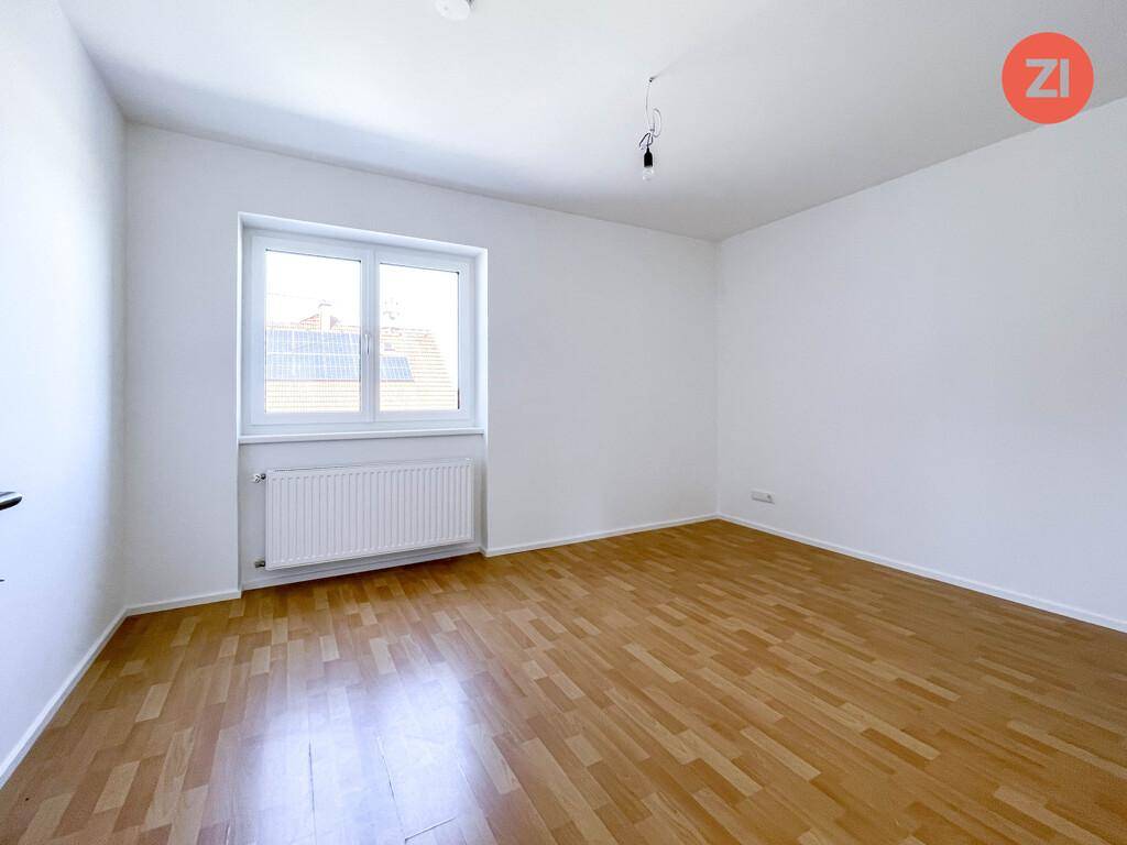 Leeres Zimmer mit Parkettboden, weiß gestrichenen Wänden und einem Fenster mit Blick auf Dächer.